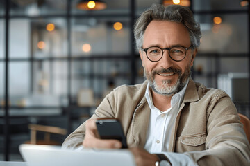 Smiling middle aged Hispanic business manager ceo using mobile phone at desk in modern office, mature Latin or Indian businessman working on smartphone and laptop