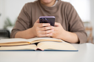 A cropped image of a female college student using her smartphone while reading a book.