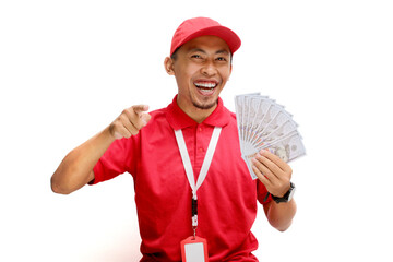 Asian delivery man or courier points towards the camera while holding money banknotes, isolated on white background. Concepts of professionalism, customer engagement, and secure monetary transactions.