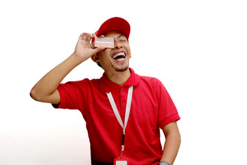 An excited Asian delivery man or courier playfully covers half of his face with a credit card, isolated on a white background. Concepts of online transactions, secure payments, customer satisfaction