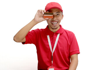 An excited Asian delivery man or courier playfully covers half of his face with a credit card, isolated on a white background. Concepts of online transactions, secure payments, customer satisfaction