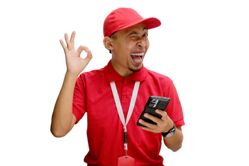 Asian delivery man or courier smiling and showing the OK gesture and winking to the camera while holding a phone. Isolated on a white background