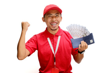 Excited Asian delivery man or courier raises his fist in triumphant YES gesture while holding money banknotes in his hand, isolated on white background. Concepts of victory, financial gains, triumphs