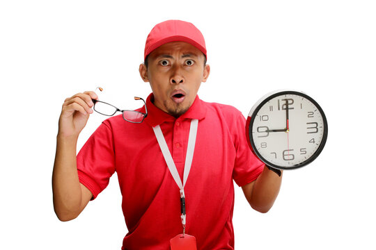 Asian delivery man or courier looks surprised as he points at a clock in his hand, isolated on a white background. Concept of unexpected deadlines, urgent deliveries, logistics and delivery services