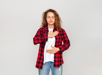 Curly brunette woman in red plaid shirt stands in front of plain wall and doing breathing exercise.