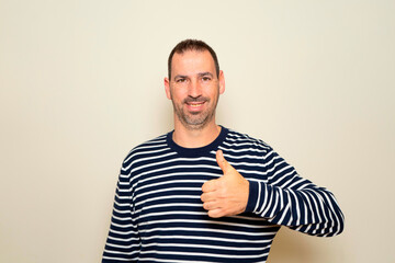 Fototapeta premium Hispanic man with beard in his 40s standing over isolated beige background doing a happy thumbs up gesture with hand. Approval expression looking at camera showing success.