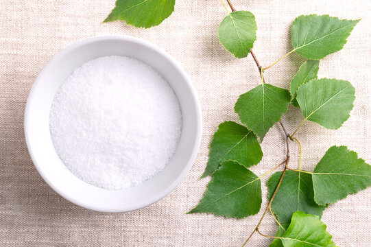 Birch sugar, table sweetener based on xylitol, in a white bowl on linen fabric. Also called xylite, a sugar substitute of natural origin with 40 percent fewer calories. With birch branch on the left.