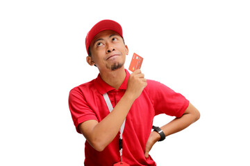 Pensive Asian delivery man or courier gazes thoughtfully at empty copy space while holding a credit card, isolated on a white background. Concept of financial planning, transactions, delivery services