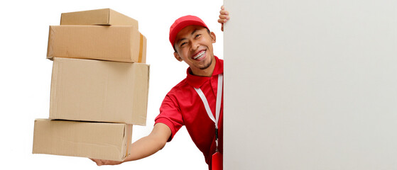 Excited Asian delivery man or courier peeks out from behind a white wall with copy space, holding a cardboard box, isolated on a white background. Delivery services, shipping, or e-commerce businesses