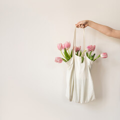 Person's hand holding a white shopping bag filled with vibrant pink tulips on light colored background. Elegant and stylish composition with delicate, soft pastel tones of the flowers and the bag