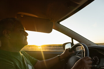 Naklejka premium Adult man driving a car during golden hour, holds the steering wheel with confidence and security on a road trip on vacation during sunset