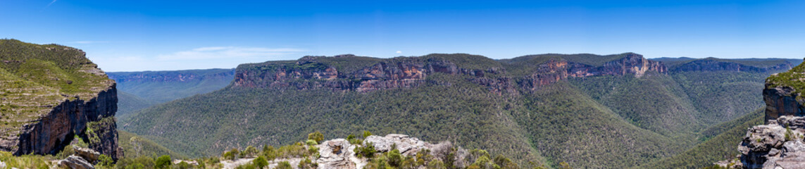 Walls Lookout, a panoramic view of the Blue Mountains National Park in New South Wales, Australia, featuring a lush valley, rugged cliffs, and a clear blue sky.
