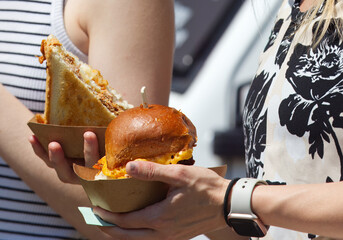 Midsection body of woman holding paper bowls with sandwich and burger at farmers street food market, low angle of view, closeup.