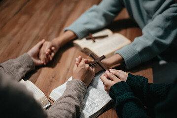Group of Christians sit together and pray around a wooden table with blurred open Bible pages in their homeroom.