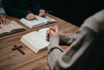 Group of Christians sit together and pray around a wooden table with blurred open Bible pages in their homeroom.