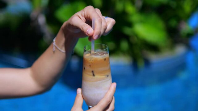 Close-up woman hand stirring iced latte with straw in glass. Refreshing iced latte against backdrop blue pool. Iced latte perfect drink for hot day by water. Summer enjoyment with cool coffee cocktail