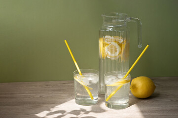 Fototapeta premium Two glasses and jug with lemonade drink and drinking straws, lemon on table with natural floral sunlight shadows, soft green background with empty copy space, summer beverage lifestyle concept