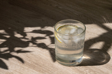 Glass with iced water with lemon slice, infused water drink, healthy summer drink for wellbeing and wellness, neutral light brown wooden table background with floral sunlight shadows