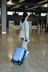 Traveler with luggage suitcase in the airport lobby before business trip. Woman with blue suitcase and phone is walking to the reception desk in the airport before flight departure.