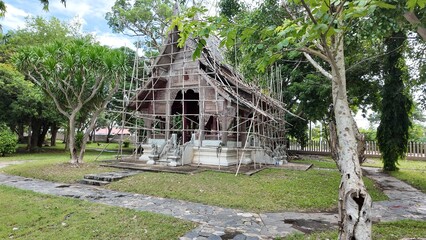 Traditional,  old Thai pavilion, surrounded by scaffolding.  Trees and bushes, a sign with Thai...