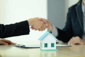 Small house model on wooden table with real estate agent shaking hands with his customer in background