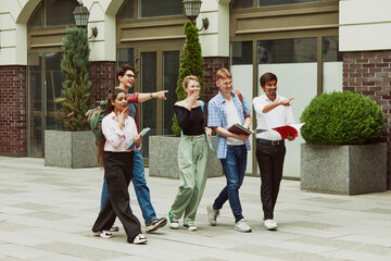 Fototapeta premium Group of multiracial people, boys and girls, students walking outside the university and pointing somewhere with excitement. Concept of education, university, multicultural students, unity