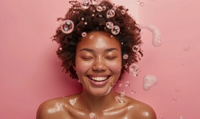 Portrait of a joyful young woman with natural curly hair enjoying a shower. She's smiling and surrounded by water drops and soap bubbles, against a pink background. Fresh and carefree moment.