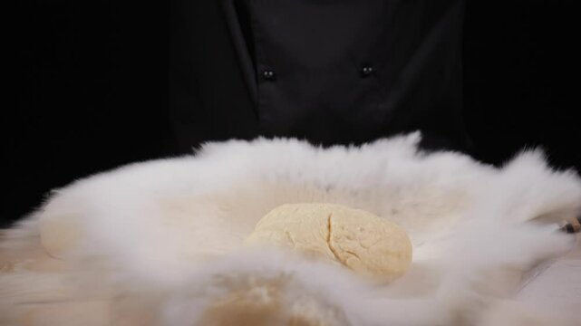 The cook prepares dough on a work surface, rolling it with a rolling pin. There is a lump of dough on the table and scattered flour, creating an effect of flying particles