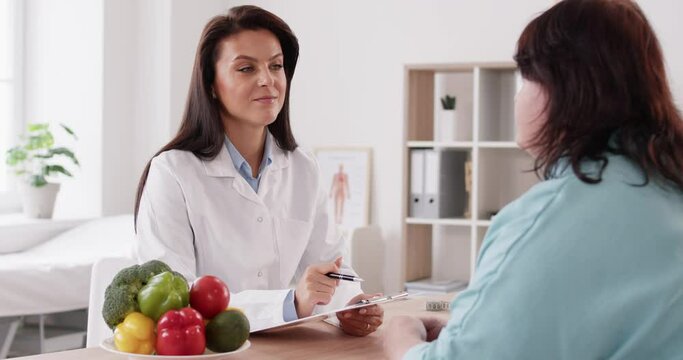 Dietician nutritionist, sitting at desk with veggies, takes notes as she listens to fat chubby woman with body weight problem asking for diet consultation, advice, recommendation, dietary prescription