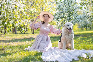 Portrait of a young woman dressed in a pink dress with a train and a hat, sitting next to a large white fluffy dog ​​against the backdrop of a spring park and blooming flowers.