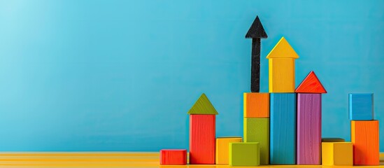 Colorful toy block towers of varying heights displayed on a yellow surface against a blue wall, with a black arrow pointing upwards in the copy space image.