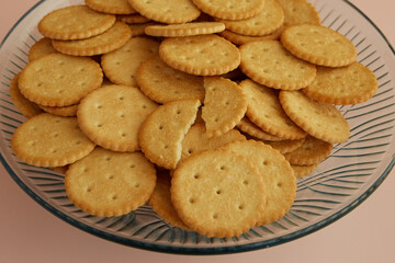 Saltine crackers in a transparent dish one of which is broken in half on a pink background close-up, cookies in a vase, cookies for tea