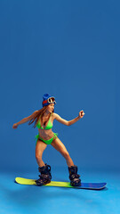 Obraz premium Stylish woman in bikini and winter sports gear, demonstrating her adventurous spirit and love for snowboarding against blue studio background. Concept of vacation, tourism, active lifestyle, fashion