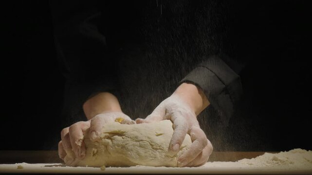 The cook kneads the dough on a flour-dusted work surface. Flour is sprinkled over the dough, creating a culinary process atmosphere. The cook's hands are covered in flour, emphasizing the preparation
