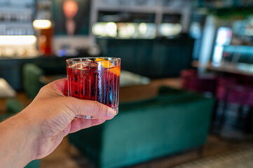 Hand holding a red cocktail with an orange slice, against a blurred bar background