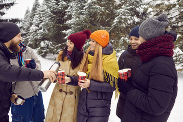 Happy friends with hot drinks in paper cups chatting and having fun outdoors in winter park. Man...