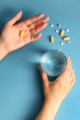Pill in one hand and glass of water in the other on a blue background with other drugs. Concept of...