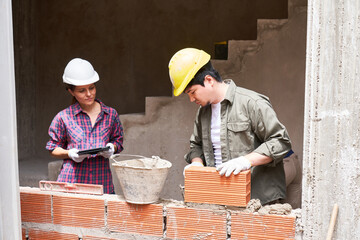 Young female contractor supervising male mason doing brickwork on wall at incomplete construction...