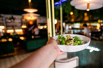 A hand holds a white plate with a green salad, sharply focused against a blurred restaurant background