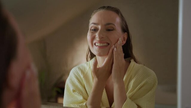 A woman stands in the bathroom, looking in the mirror while experiencing tooth pain. She is touching her jaw and face, grimacing in discomfort