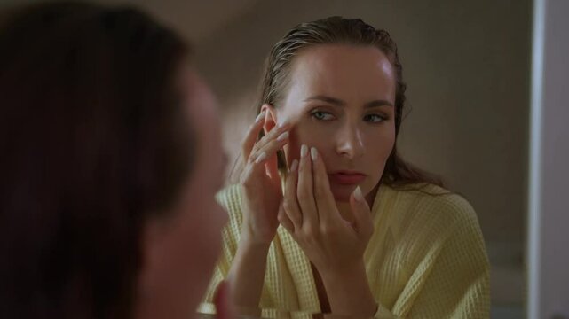 Woman looking in a mirror, focusing on the pimples on her face. She looks concerned and worried about her skin