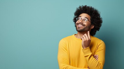 Obraz premium A cheerful man with curly hair and glasses in a bright yellow sweater, smiling and looking upwards against a teal background. Ideal for themes of thinking, optimism, and creativity