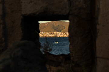 View of a boat in the sea through a small hole window in an old brick wall. A city and a mountain in the background.