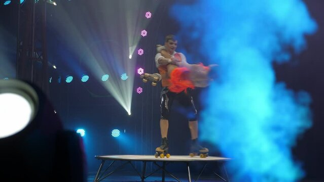 A pair of acrobats on roller skates perform a dangerous trick in the circus arena, the performance of gymnasts on roller skates under the light of colorful spotlights and smoke. A circus show.