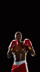 Portrait of focused African man with shirtless strong muscular body, boxer in gloves and shorts, standing in stance isolated on black background. Concept of sport, body, strength. Vertical poster