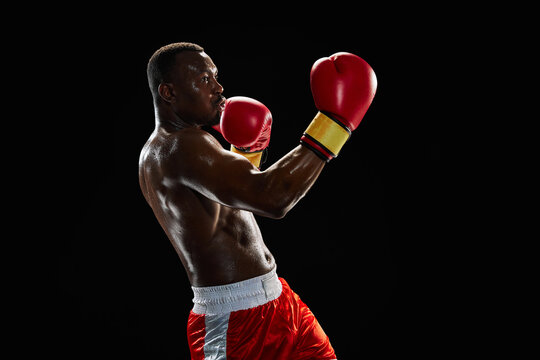 Uppercut punch. Motivated and ambitious sportsman, muscular boxer in motion fighting, training shirtless in gloves isolated on black background. Concept of sport, active lifestyle, body, strength