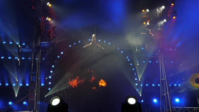 A circus show with tightrope walkers under the dome of the circus under the light of colorful spotlights. An acrobat girl sits on a rope splits under the dome of the circus.