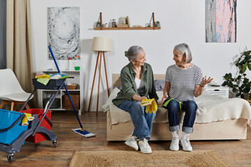 A mature lesbian couple enjoys a break from housework, relaxing on their bed and chatting in their modern apartment.