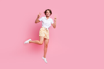 Full body photo of jumping young girl wear yellow cute shorts combination with glamour blouse show v-sign isolated on pink color background