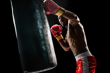 Strong man, boxing athlete intensely training with punching bag, demonstrating strength and determination isolated on black background. Concept of professional sport, active lifestyle, body, strength